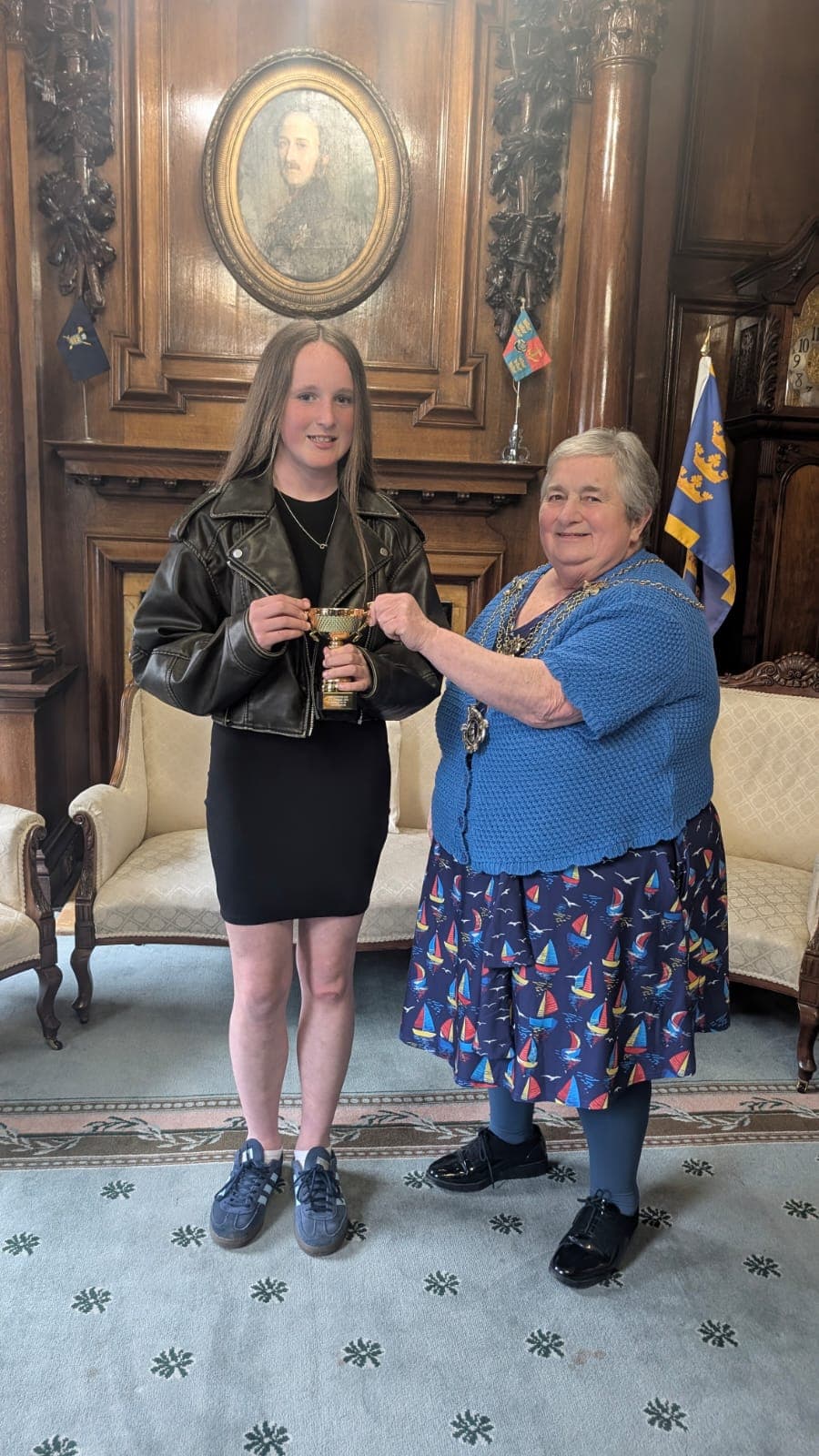 Two women in formal room, one handing a trophy to the other