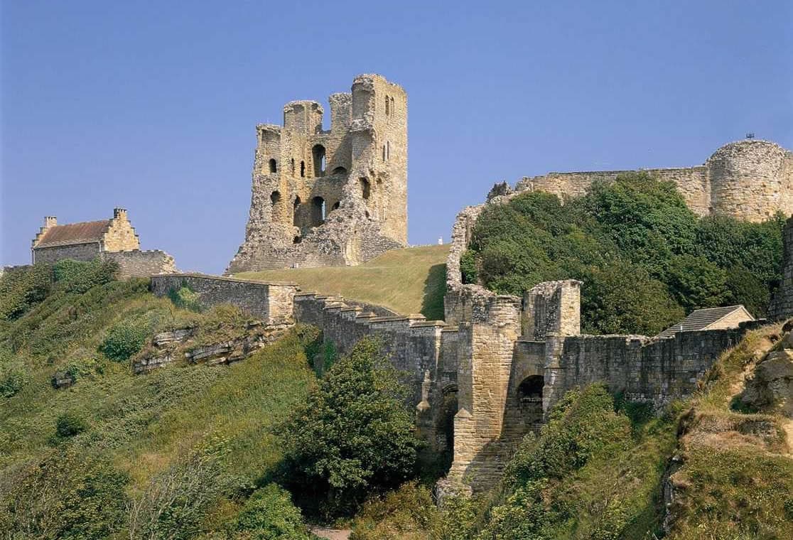 Ruins of Scarborough Castle stand against a blue sky, overlooking the sea and green landscape.