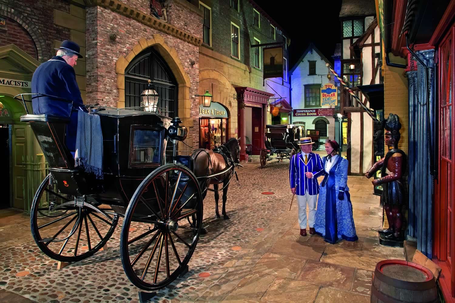Victorian street scene with horse-drawn carriage, cobblestone path, and people in period costumes outside colorful shops.