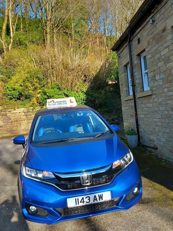 Blue car parked near a stone building, surrounded by greenery and trees in a sunny setting.