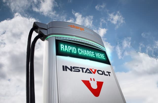 InstaVolt charging station with a digital display reading "RAPID CHARGE HERE" against a blue sky with clouds.
