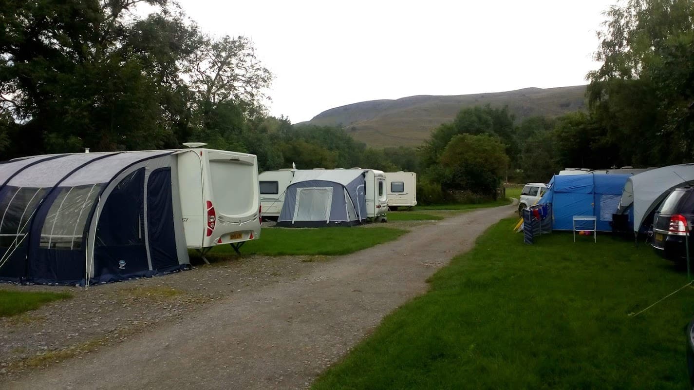 Campsite with tents and caravans on a grassy area, surrounded by trees and hills in the background.