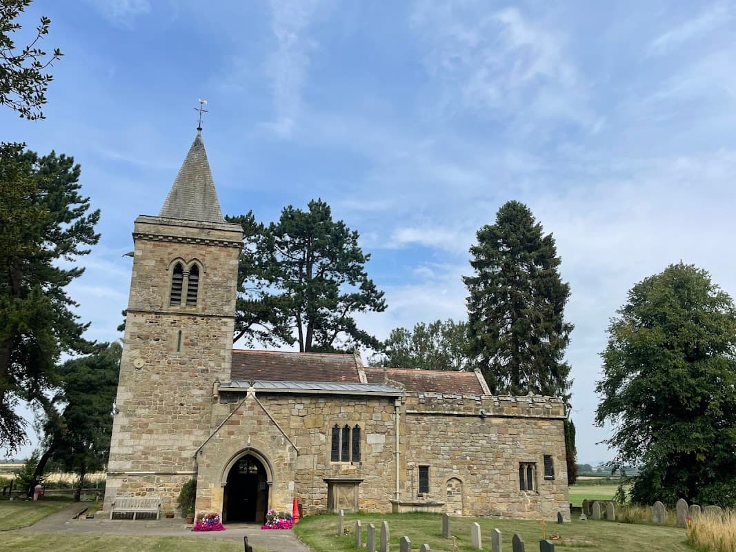 Historic stone church with a tall steeple, surrounded by trees and a grassy graveyard under a blue sky.