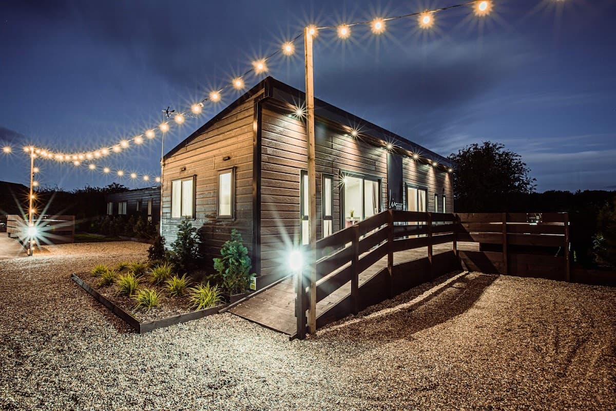 Modern wooden building with outdoor lighting, surrounded by gravel and greenery, set against a twilight sky.