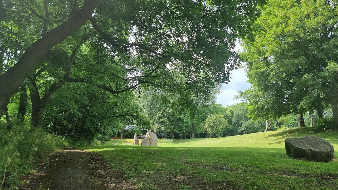 Lush green park with large trees, a grassy area, and stone sculptures under a partly cloudy sky.