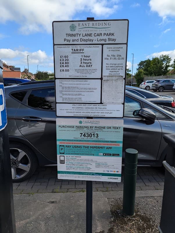 Signboard displaying parking tariffs and payment options at Trinity Lane Car Park, with cars parked nearby.
