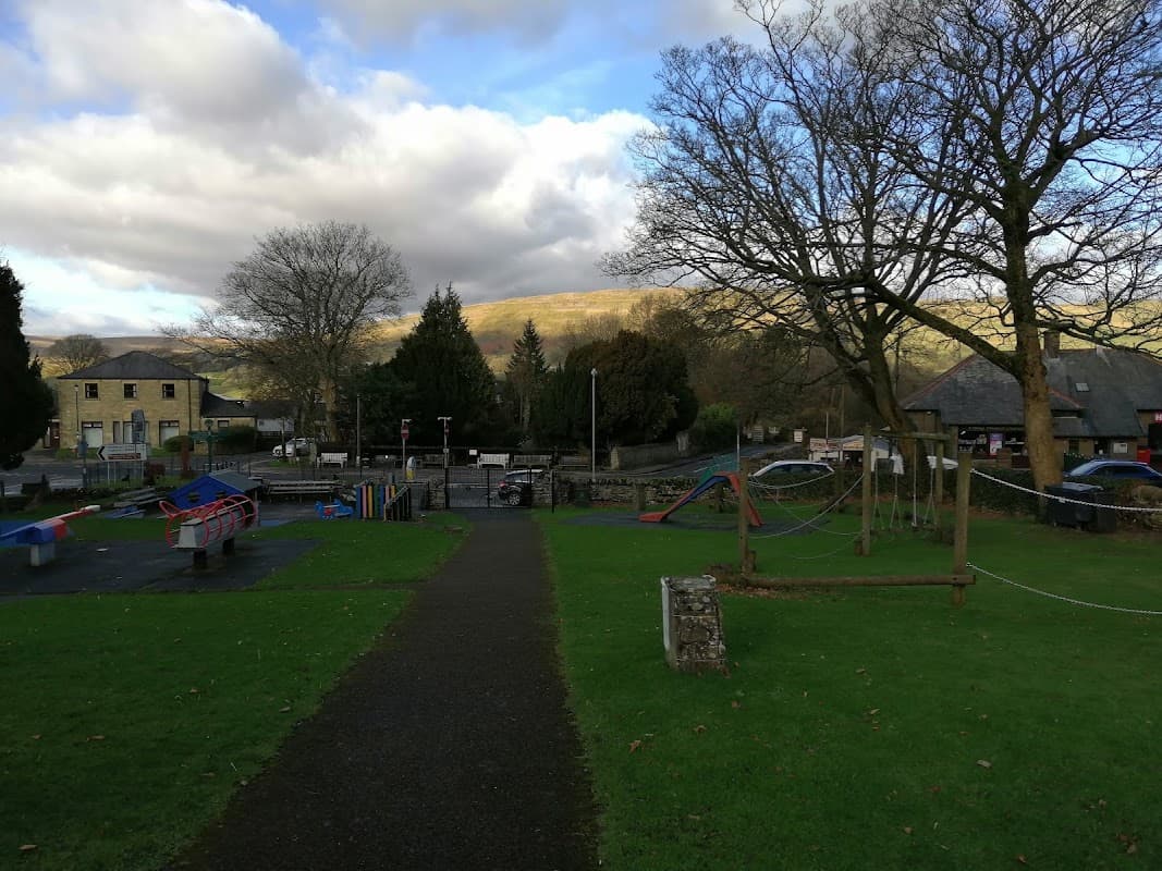 Playground with climbing structures, swings, and benches, surrounded by grass and trees under a cloudy sky in Burtersett.