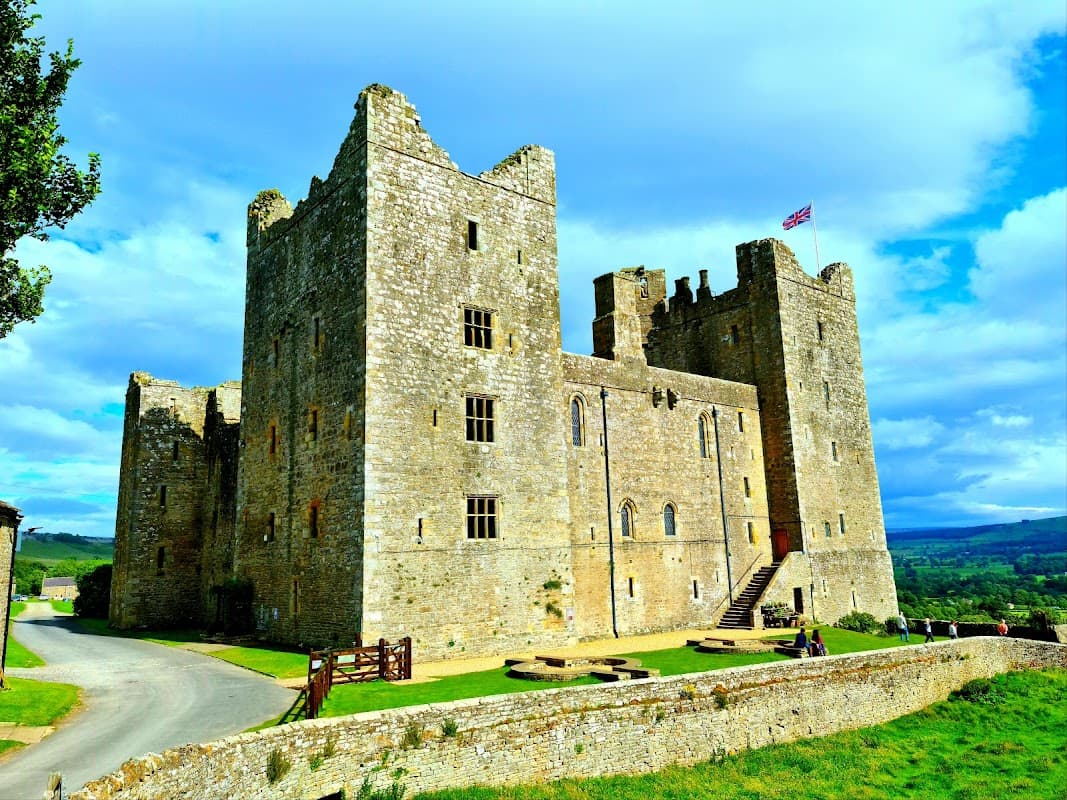 Historic Bolton Castle features stone walls, towers, a flag, and lush green surroundings under a bright blue sky.