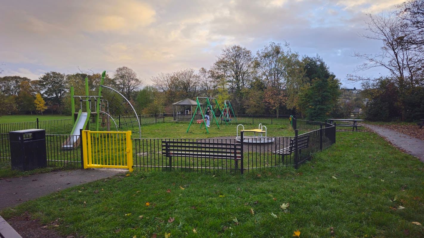 Playground with swings, slides, and a roundabout, surrounded by green grass and trees under a cloudy sky.