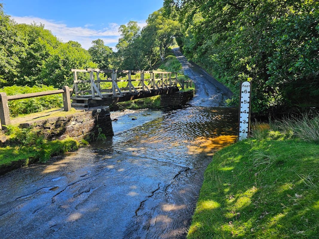 A wooden bridge over a stream with a depth marker, surrounded by lush greenery and a sloping road in Commondale.