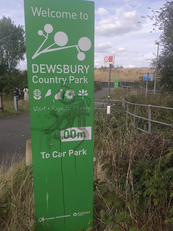 Welcome sign for Dewsbury Country Park with greenery and a path leading to the car park in the background.