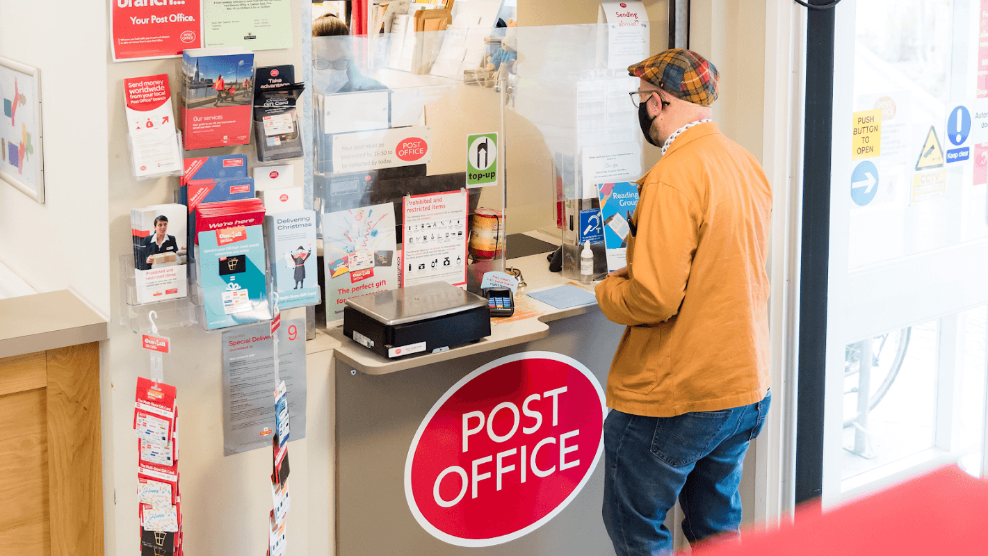 Customer at a counter in New Earswick Post Office, surrounded by brochures and a prominent red "POST OFFICE" sign.