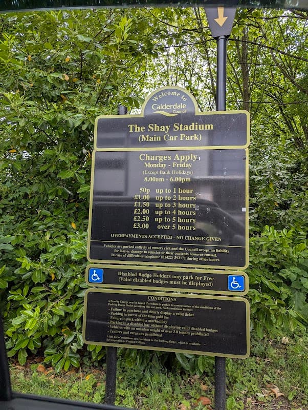 Sign detailing parking charges and conditions at Shay Stadium car park, surrounded by greenery in Exley, Yorkshire.
