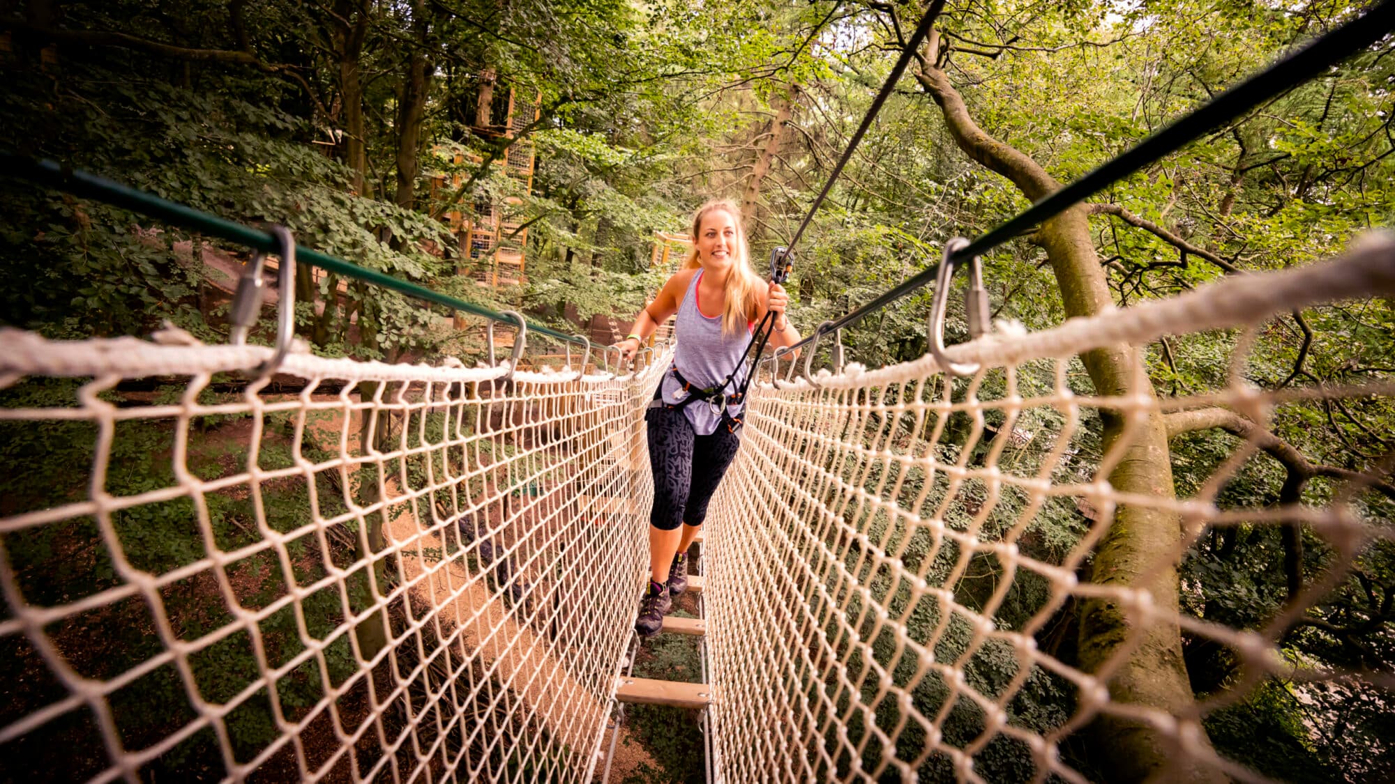 Child navigating treetop rope bridge in lush forest, with adults and other children in background, all wearing safety harnesses.