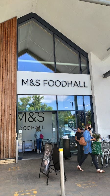 Modern storefront of M&S Foodhall with large glass entrance, wooden accents, and shoppers outside.