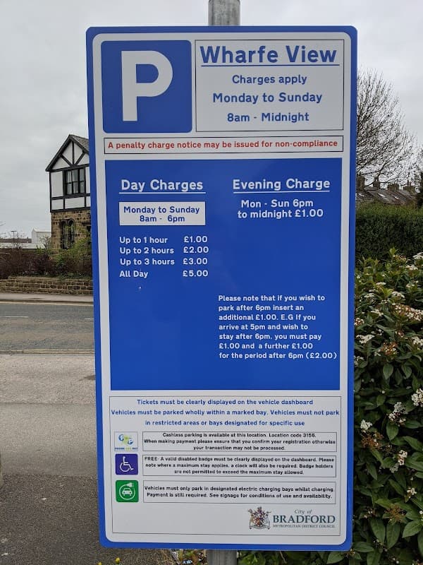 Sign for Wharfe View Car Park detailing parking charges and hours, with nearby buildings and trees in the background.