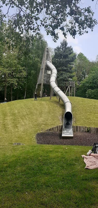 Large silver slide winding down a grassy hill, surrounded by trees and a playground area in Hemsworth Waterpark.