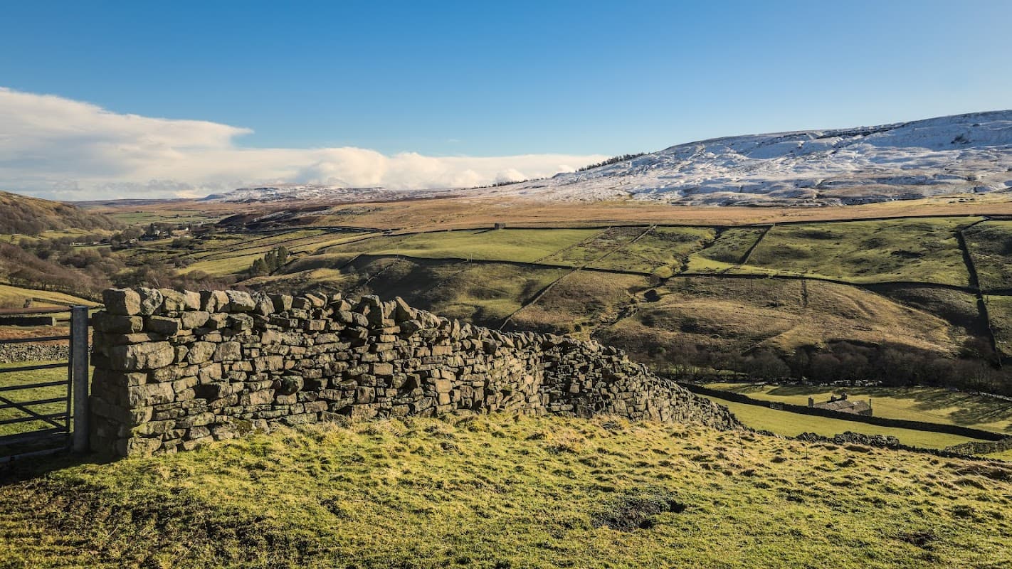Stone wall in foreground with rolling green hills and snow-capped mountains under a clear blue sky in Langthwaite, Yorkshire.