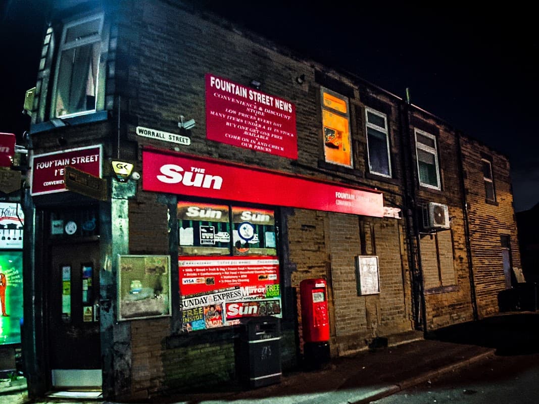 Fountain Street News corner shop at night, featuring a red sign, newspaper displays, and a vintage red phone box.