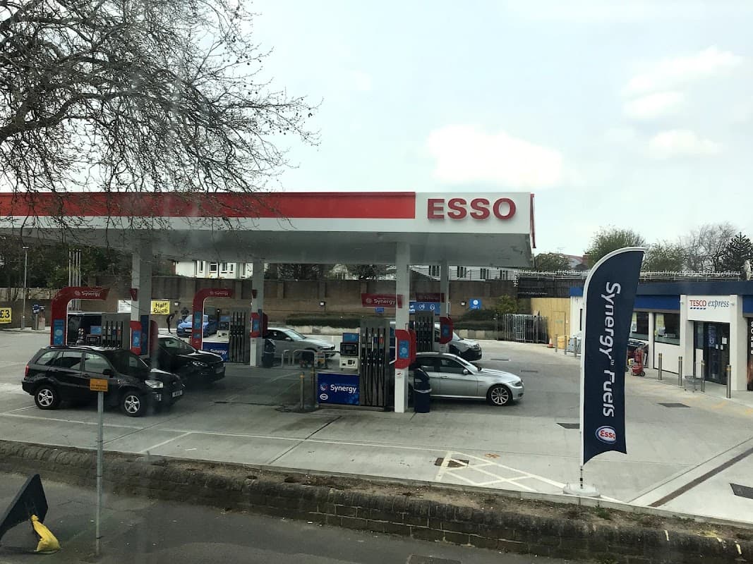 Tesco Esso Express station with multiple fuel pumps and cars, surrounded by trees and a clear sky in Leeds.