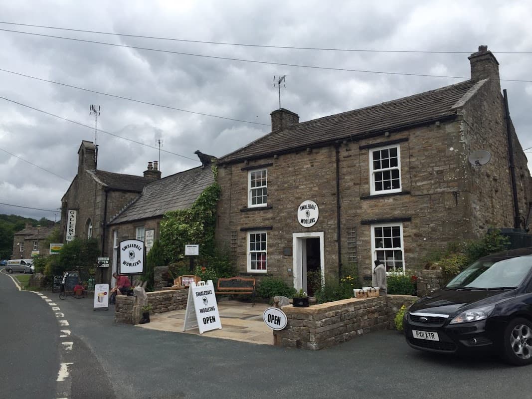 Stone shopfront with "Swaledale Woollens" sign, potted plants, open sign, and parked cars on a cloudy day in Muker.