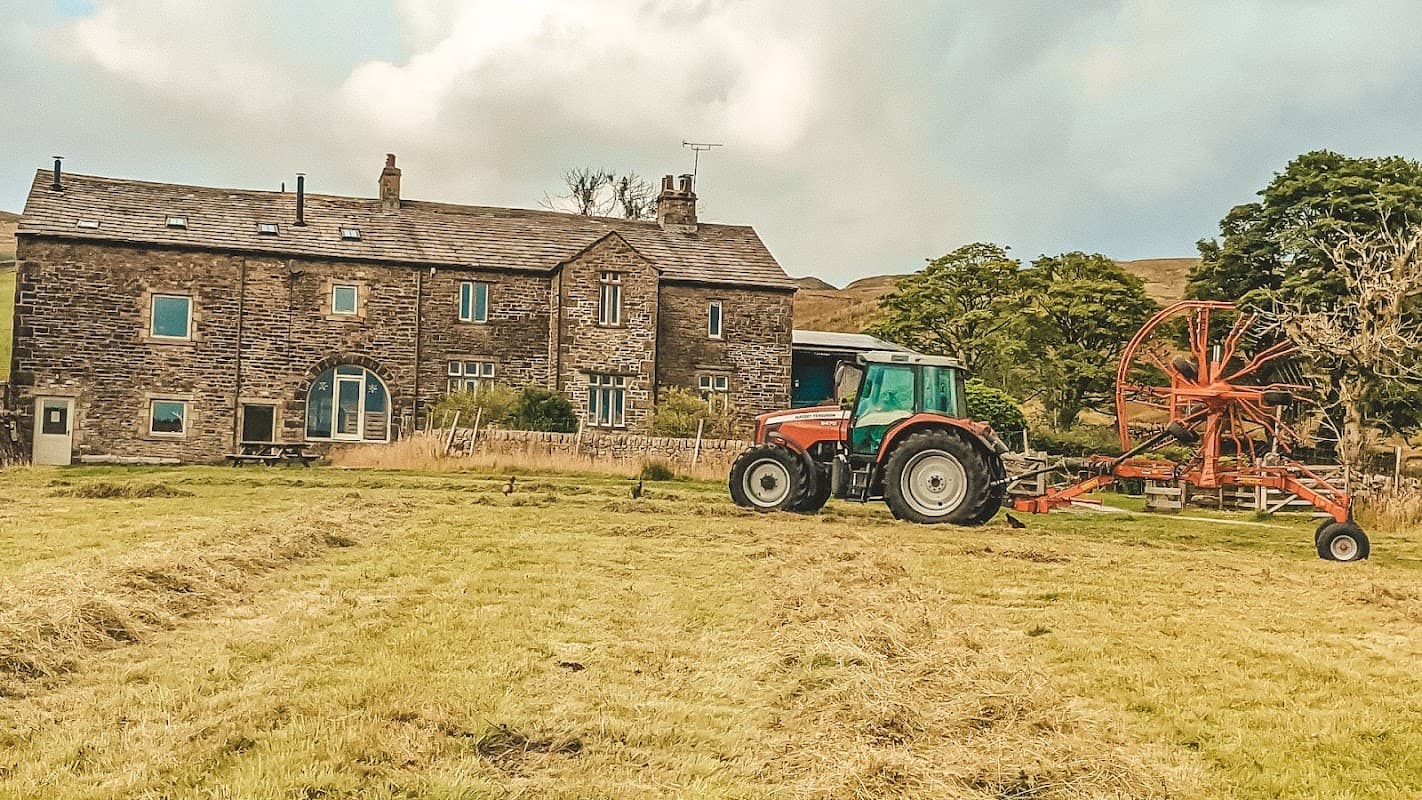 Stone farmhouse with large windows, surrounded by fields, and a red tractor with a hay rake in the foreground.
