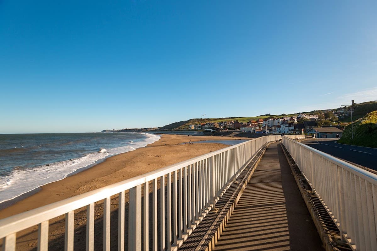 Coastal view from a walkway, with sandy beach, waves, and a quaint village in the background under a clear blue sky.