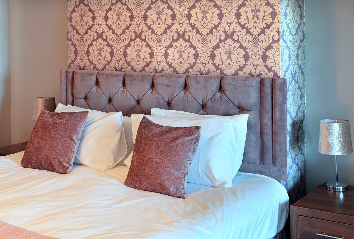 Elegant hotel room with a plush tufted headboard, white bedding, and decorative pillows against a floral wallpaper backdrop.