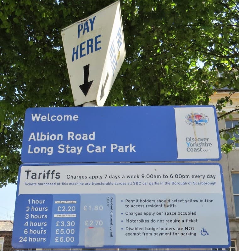 Sign for Albion Road Long Stay Car Park in Scarborough, detailing tariffs and payment instructions under a leafy tree.