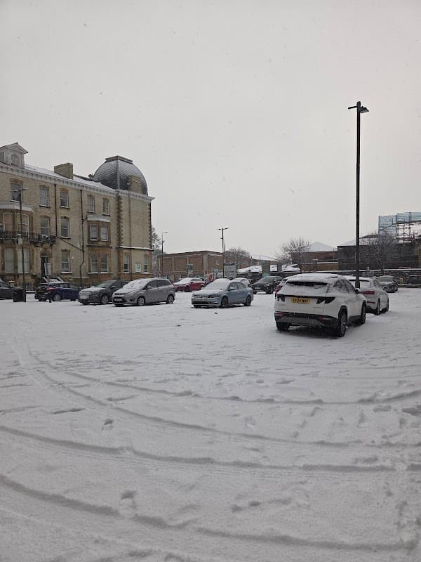 Snow-covered car park with parked vehicles and a historic building in the background under a gray sky.