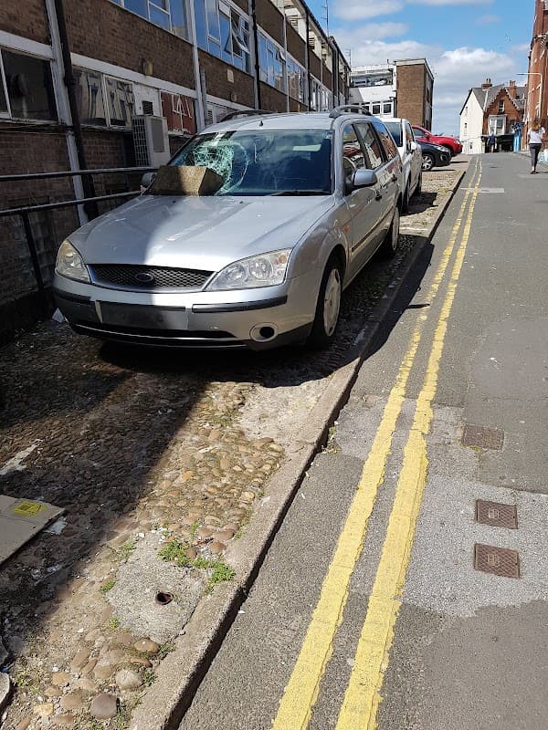 Silver car parked on a narrow street with yellow lines, surrounded by buildings and a clear blue sky.