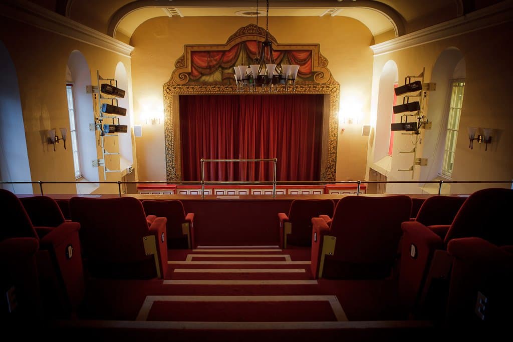 Theatrical stage with a large painted backdrop, ornate curtains, and wall sconces in a vibrant community hall.