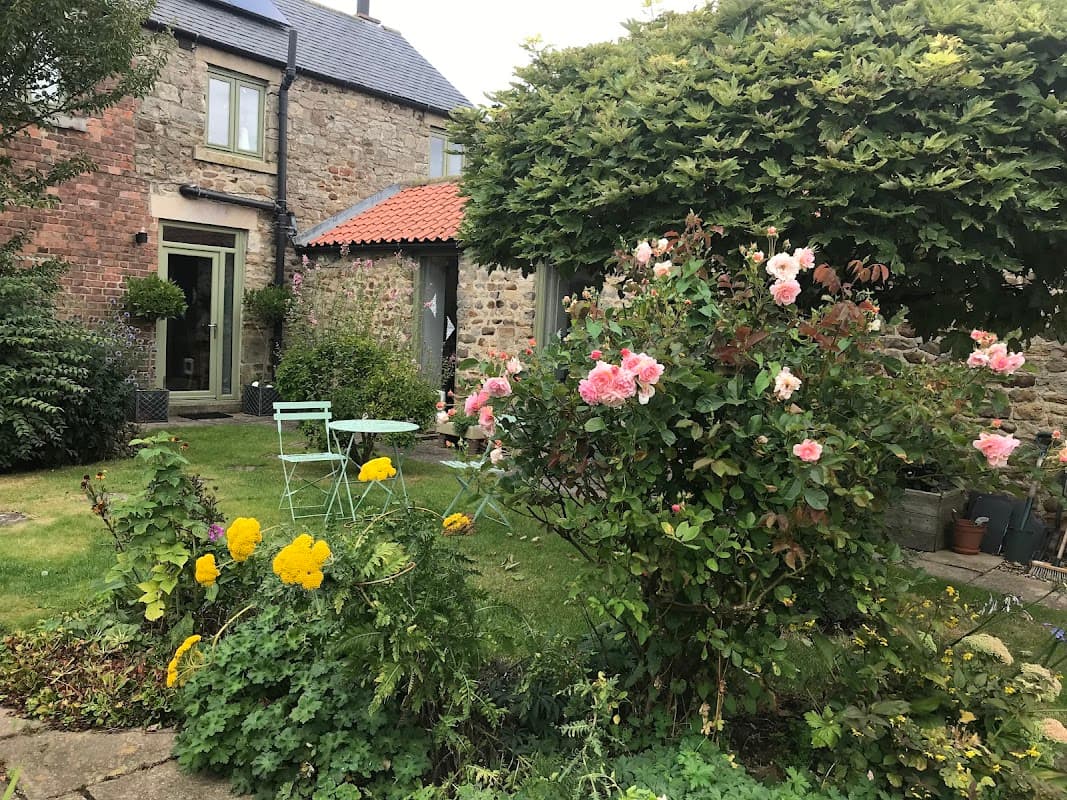 Charming guest house with stone walls, vibrant flower garden, and green seating area under a lush tree.