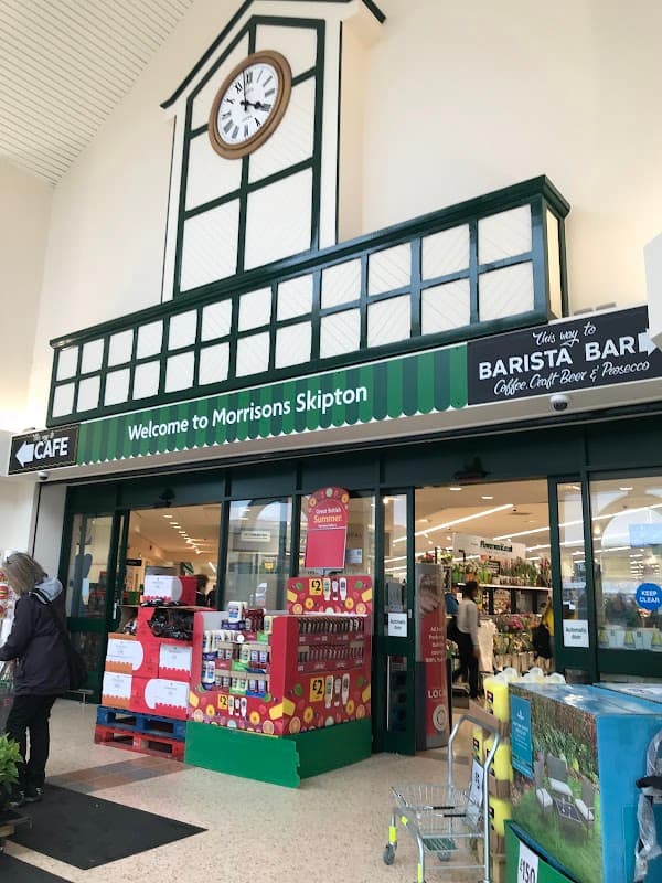 Entrance to Morrisons in Skipton, featuring a clock, café sign, and colorful displays of products outside.