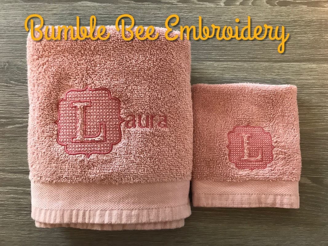 Pink towels with embroidered initials "L" and "Laura" displayed on a wooden surface, with "Bumble Bee Embroidery" text above.