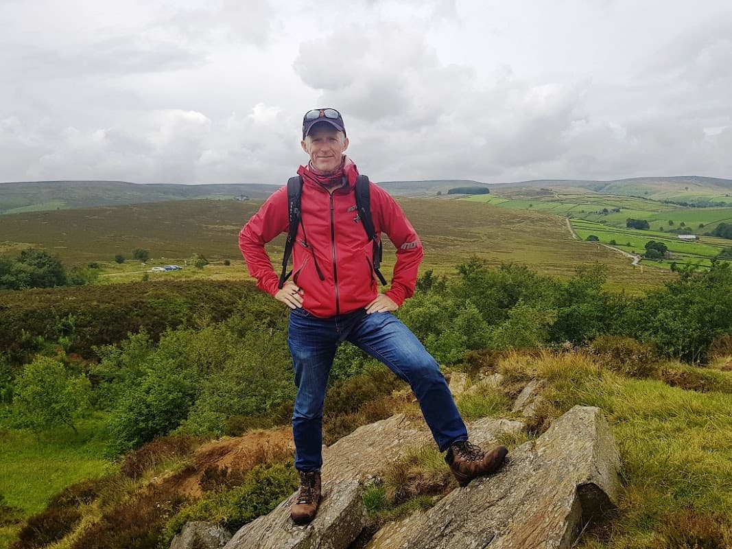 A man in a red jacket stands confidently on a rock, surrounded by lush green hills and a cloudy sky in Yorkshire.