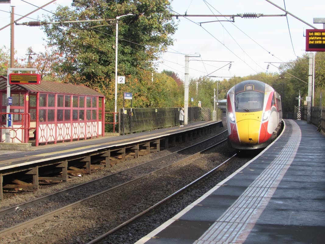 Train approaching Outwood Station, with a red and white shelter and platform signs visible. Trees in the background.