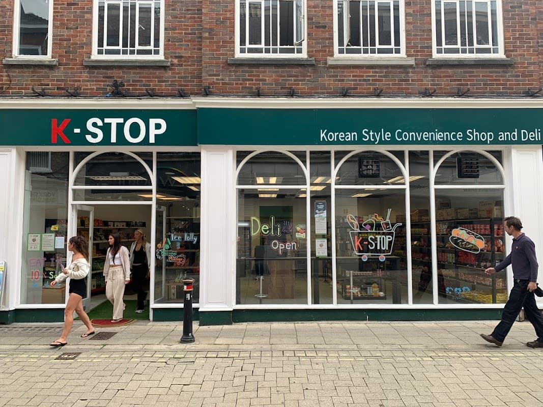 K-STOP store front with large windows, displaying colorful products and a "Deli Open" sign, people walking by.