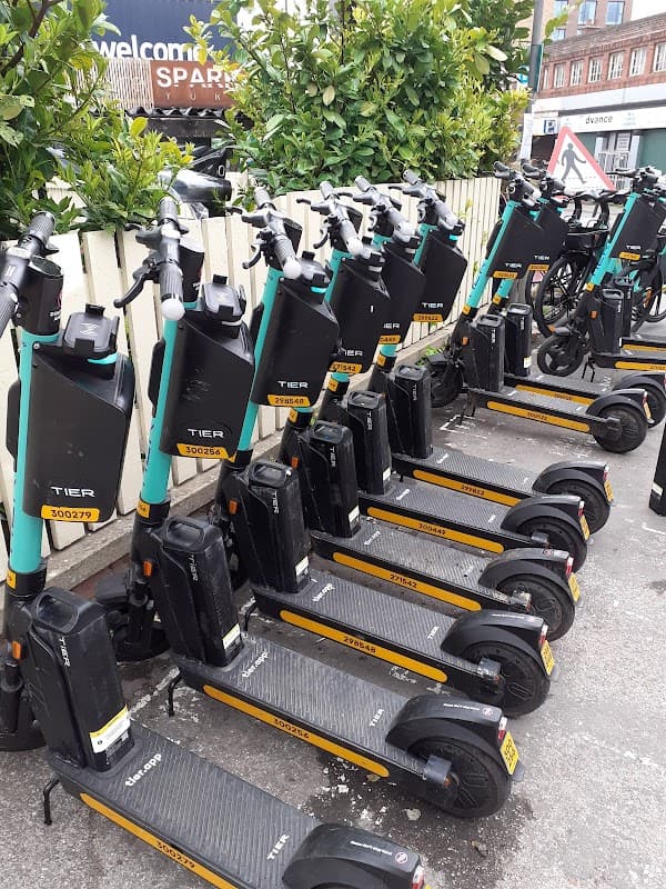 Row of electric scooters parked neatly, with greenery in the background and a sign for Spark Piccadilly visible.