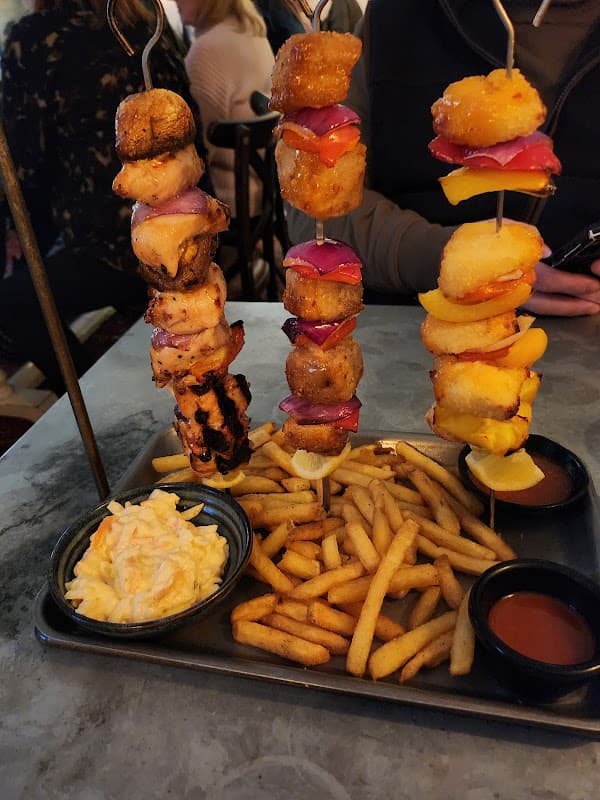 Skewers of grilled meats and vegetables served with coleslaw and fries on a metal tray, set in a busy restaurant.