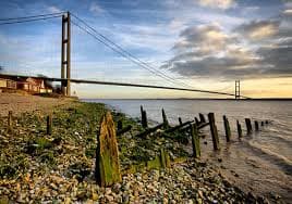 Humber Bridge spans across a cloudy sky, with a rocky shoreline and old wooden posts in Hessle, Yorkshire.