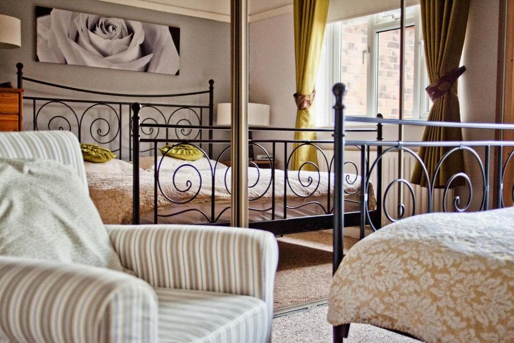 Cozy Yorkshire hotel room with iron bed frames, striped armchair, green curtains, and floral decor. Soft, inviting ambiance.