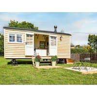 Wooden shepherd's hut with white doors, large windows, and small porch, set in a grassy area with outdoor seating.