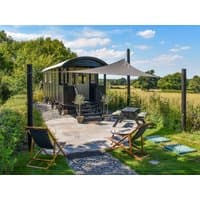 Green shepherd's hut with arched roof, patio, outdoor seating, and sunshade, set in a lush countryside landscape.