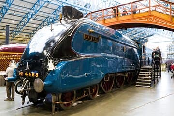 Streamlined blue steam locomotive under a glass roof with visitors nearby exploring the train exhibit.