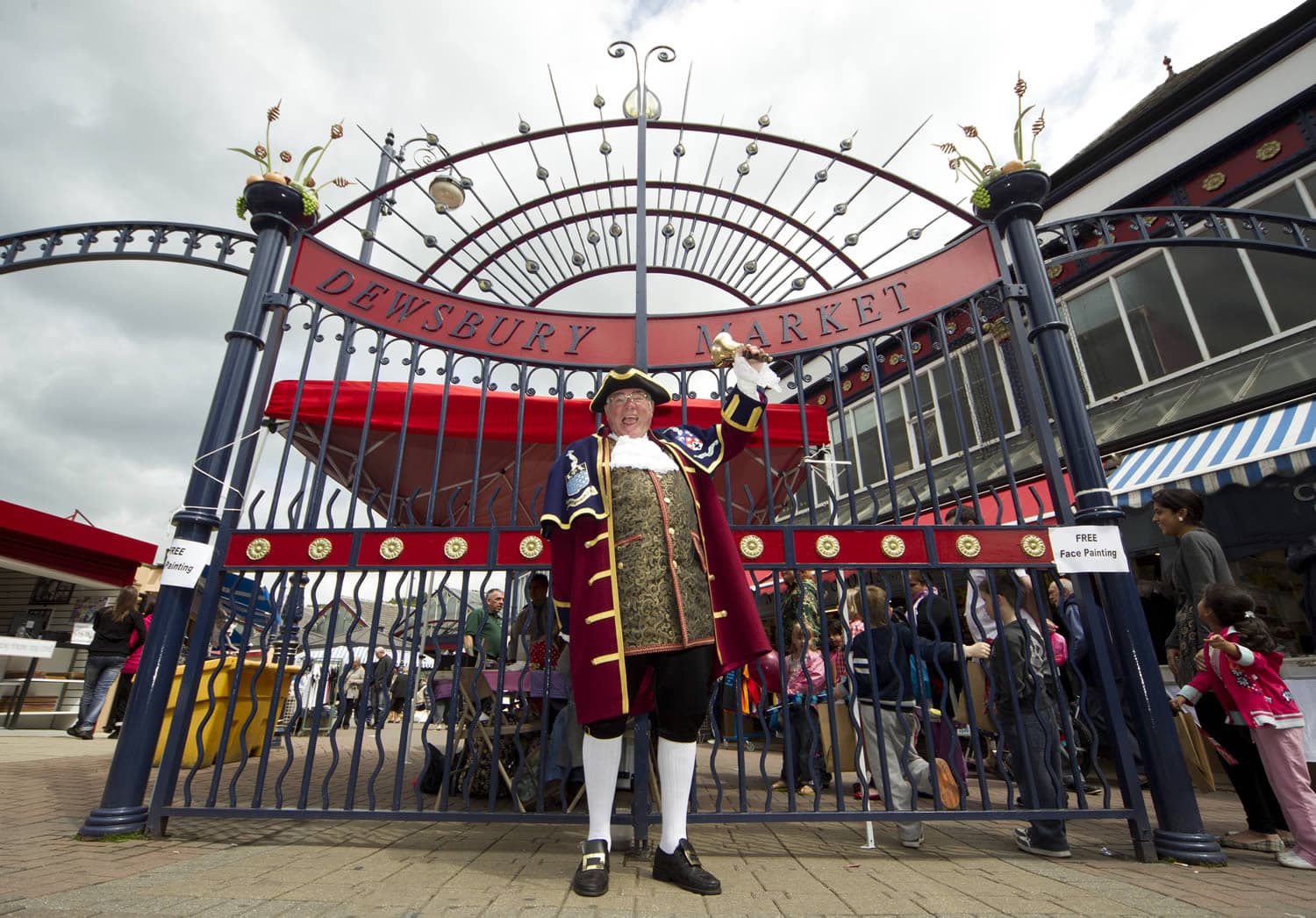 Town crier in traditional attire stands before ornate metal gate of Dewsbury Market, with bustling crowd in background.