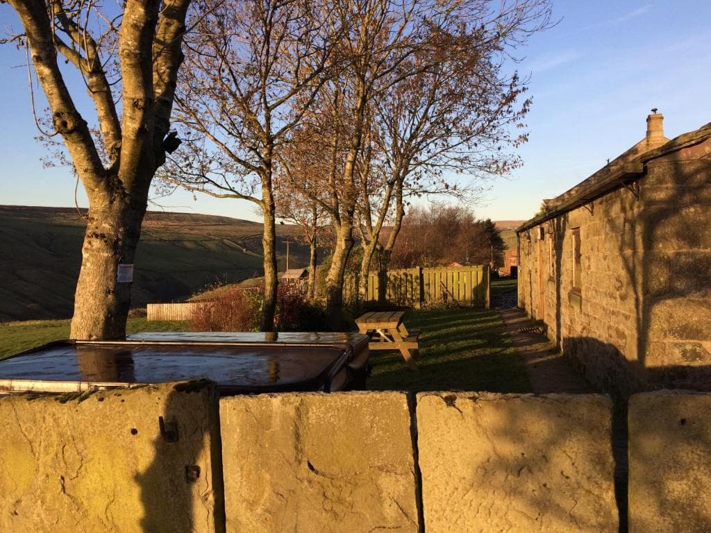 Stone cottage with a rustic roof, surrounded by bare trees, a wooden picnic table, and a scenic hillside landscape.
