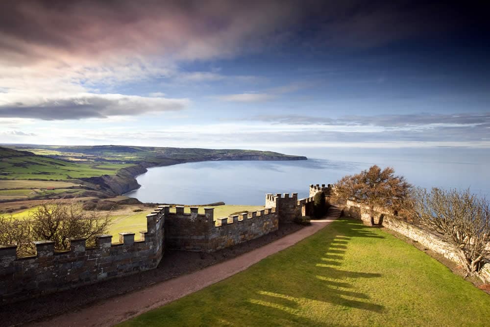 Stone battlements overlooking a green cliffside and expansive sea under a cloudy sky.