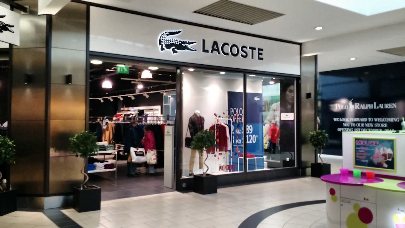 Lacoste shoe shop entrance featuring logo, window displays of apparel, and decorative plants in a shopping center.