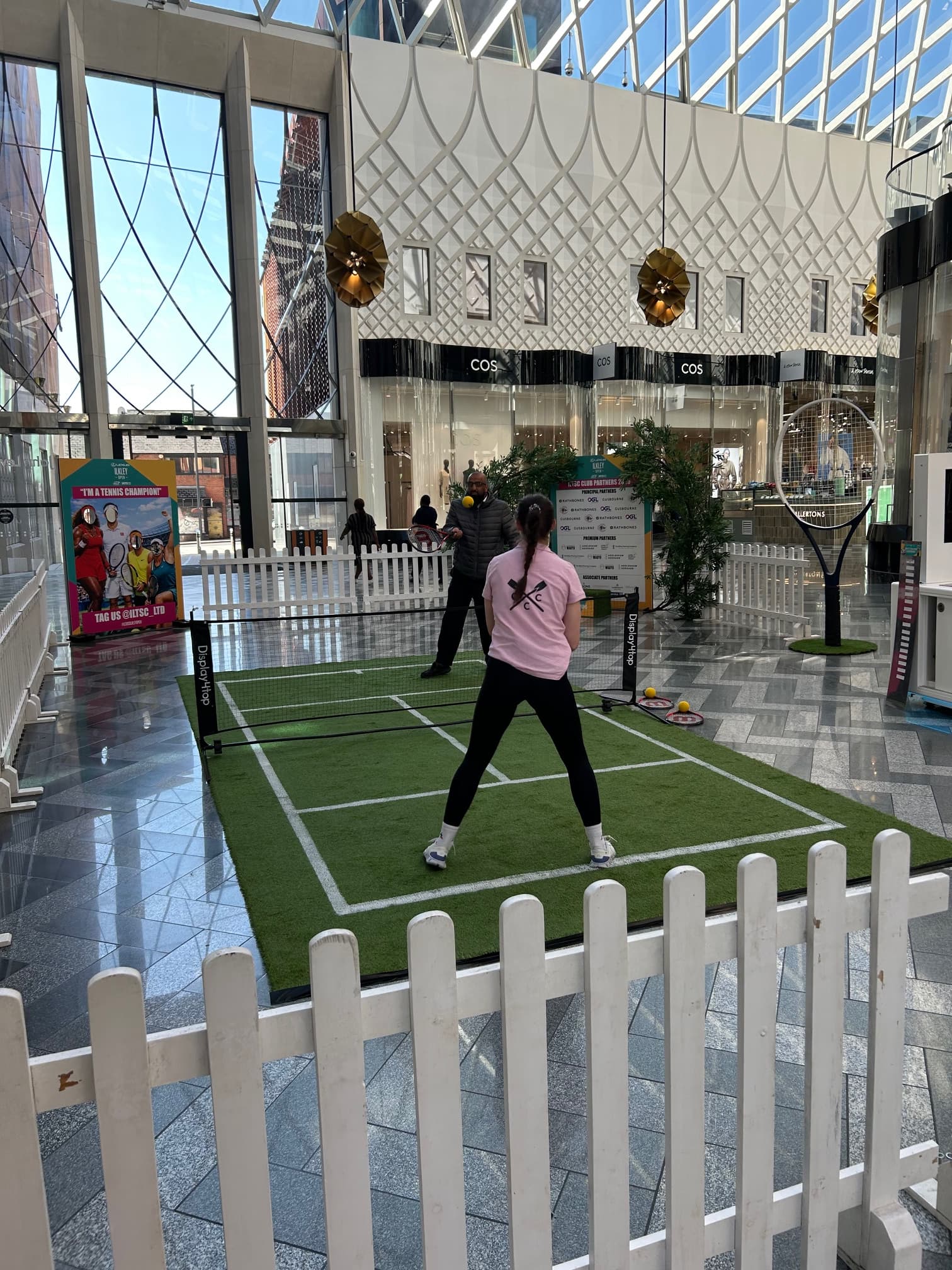 A vibrant pop-up tennis court in Victoria Gate, Leeds, with players enjoying the game inside a modern shopping center.